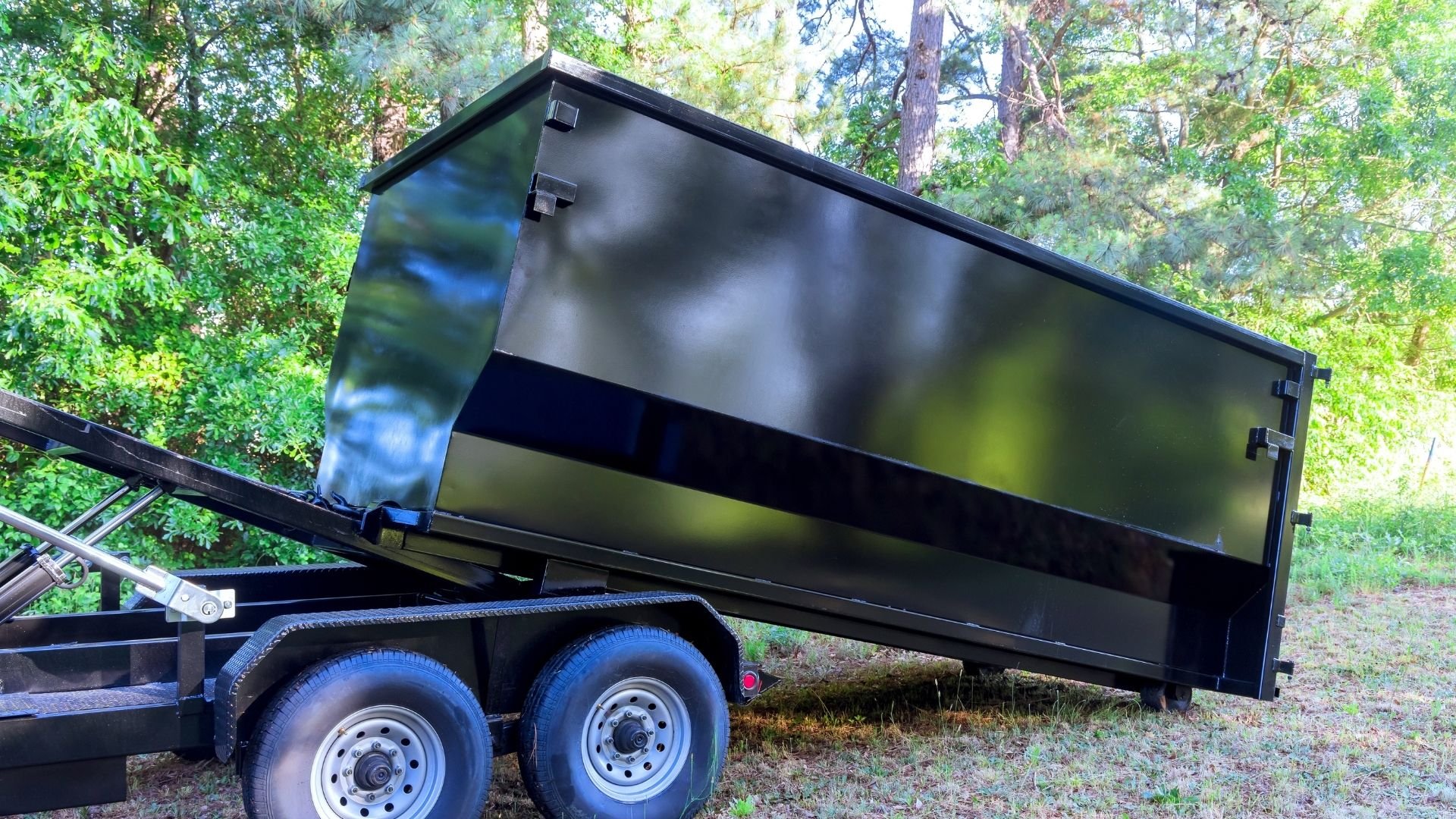 Black dumpster trailer with dual axles parked in wooded area surrounded by trees