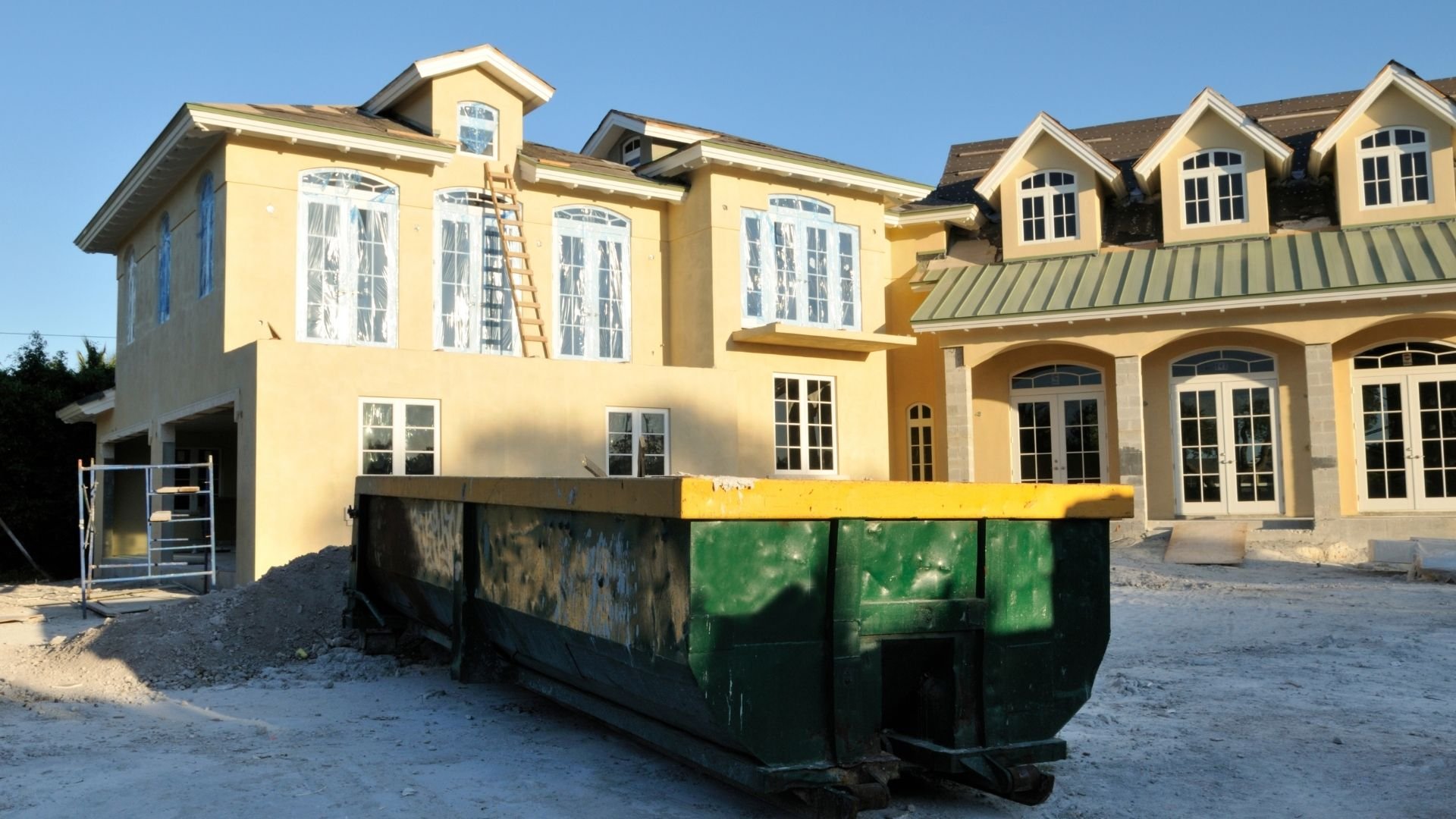 Green and yellow Dumpster in front of large yellow residential home under construction