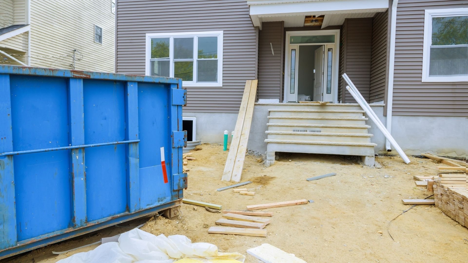 Blue dumpster on construction site with wooden debris and residential house entrance