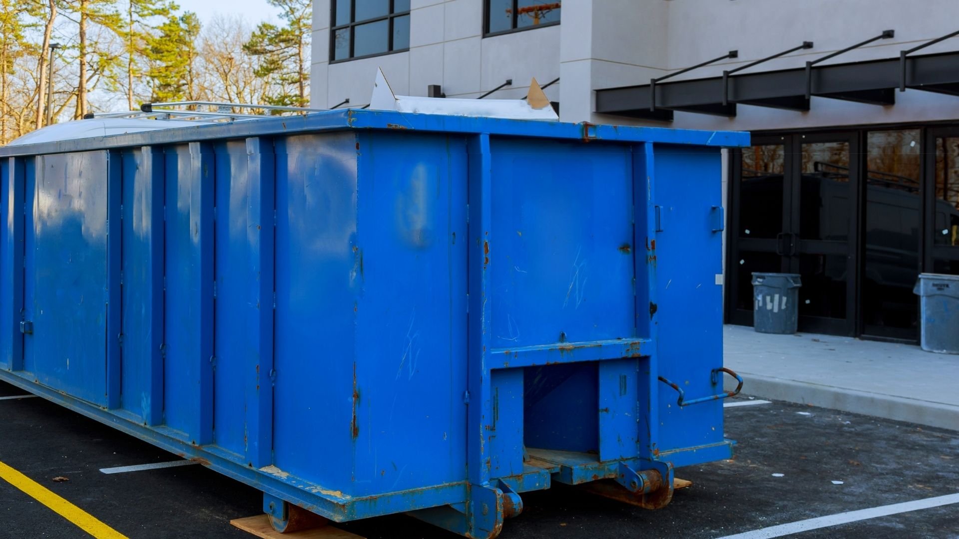 Large blue metal dumpster with doors positioned outside modern white building with trees.
