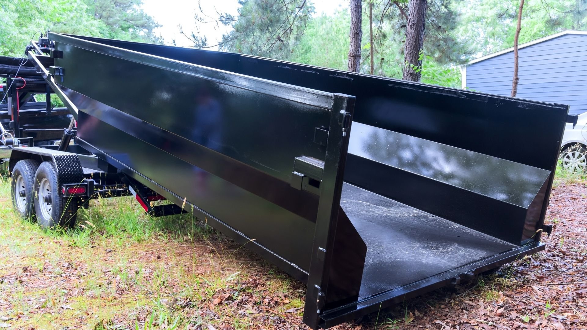 Black metal dumpster trailer parked on wooded residential property with tilted bed.